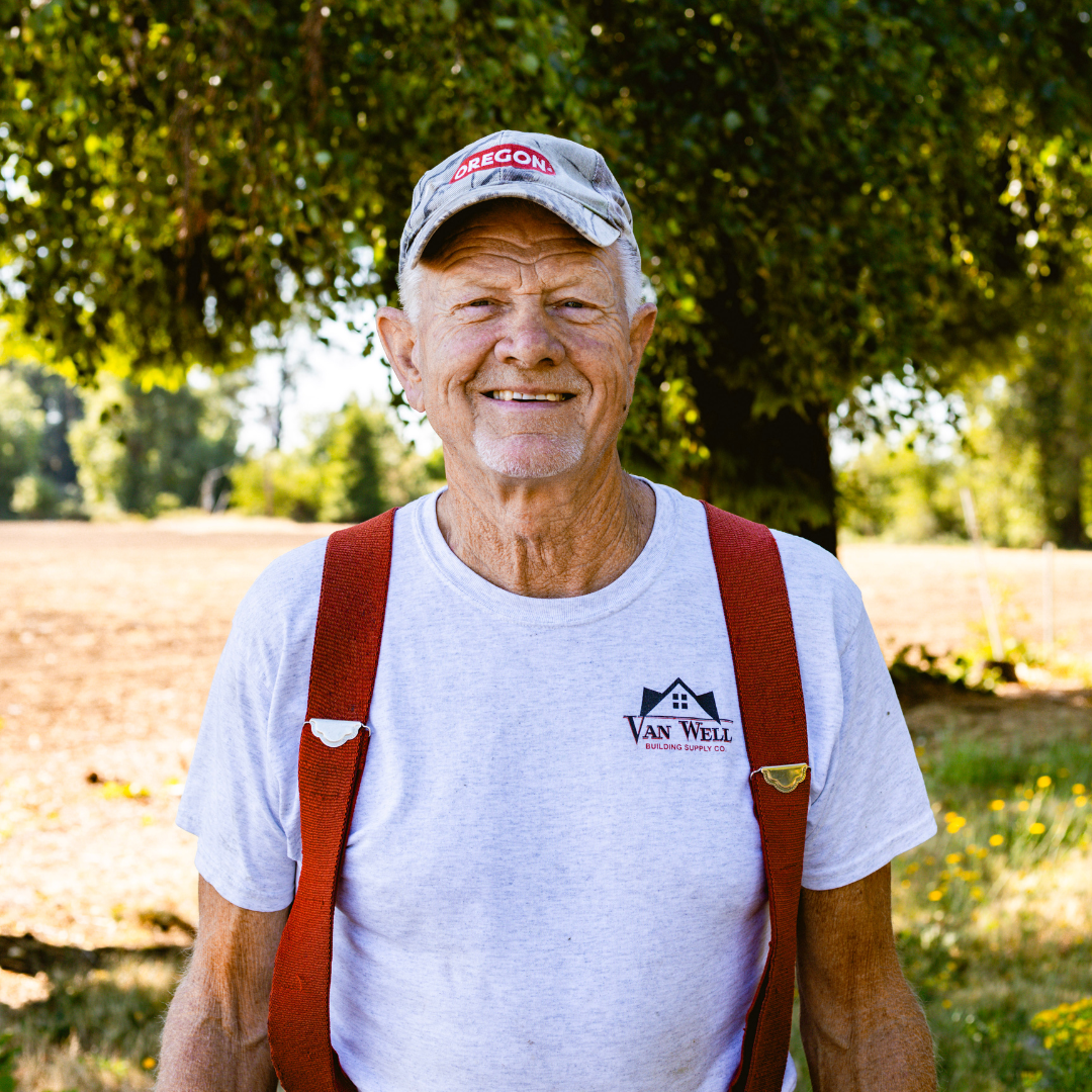 An elderly man smiling outdoors, wearing a gray baseball cap with 'Oregon' embroidered, a light gray T-shirt with a 'Van Welling' logo, and red suspenders, standing in front of a tree with green leaves and a field.