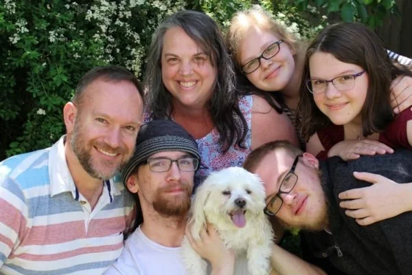 A family of six people and a small white dog posing outdoors in front of green foliage. The family members include three men, one woman, and two young girls. They are smiling and appear happy.