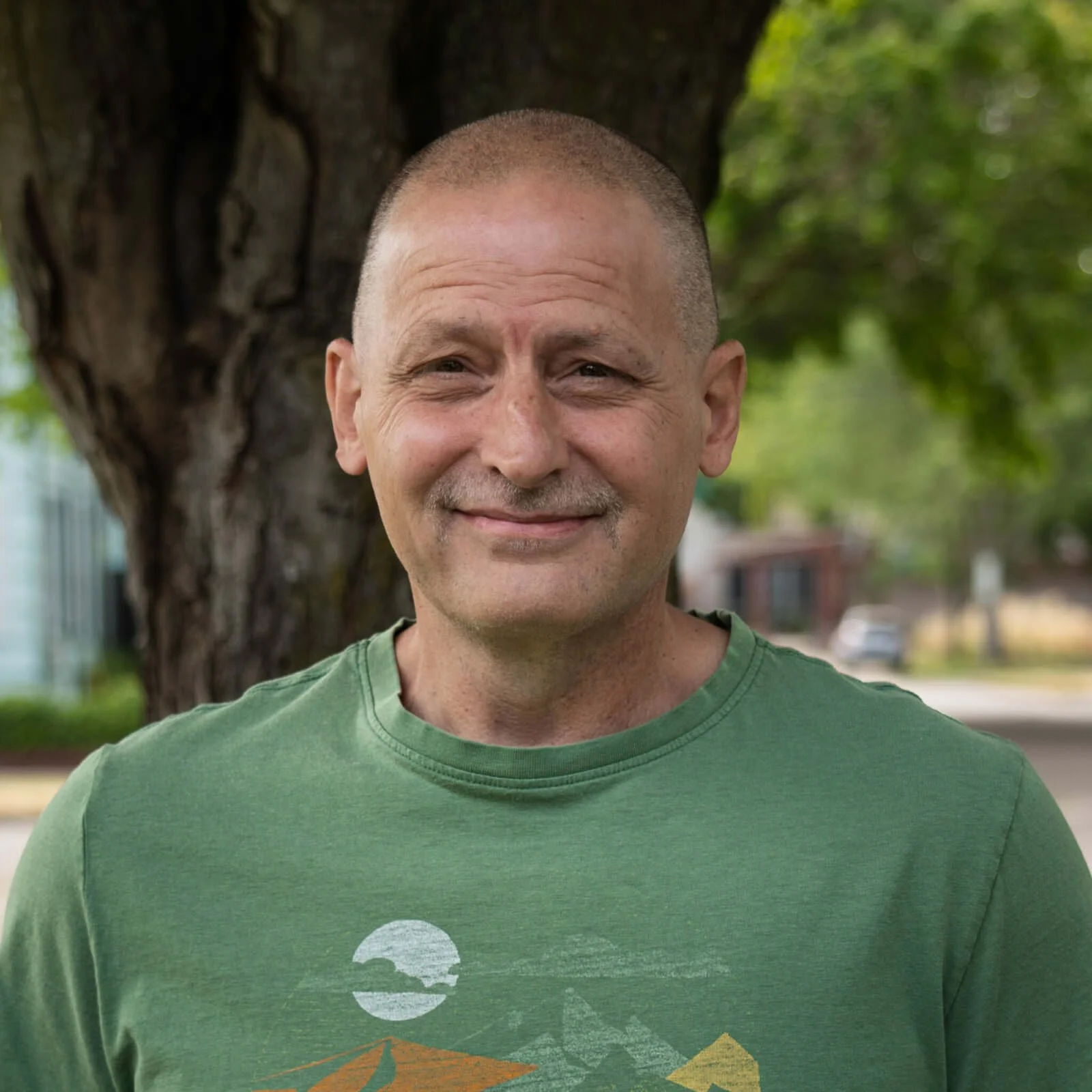 A middle-aged man with short hair and a slight smile, wearing a green T-shirt with a graphic design, standing outdoors in front of a large tree with green leaves and a blurred background of a neighborhood street.