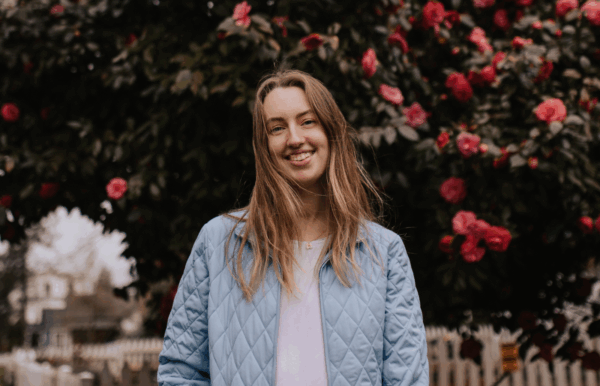Young woman with long hair smiling, wearing a light blue quilted jacket, standing outdoors in front of a flowering bush with pink blossoms.