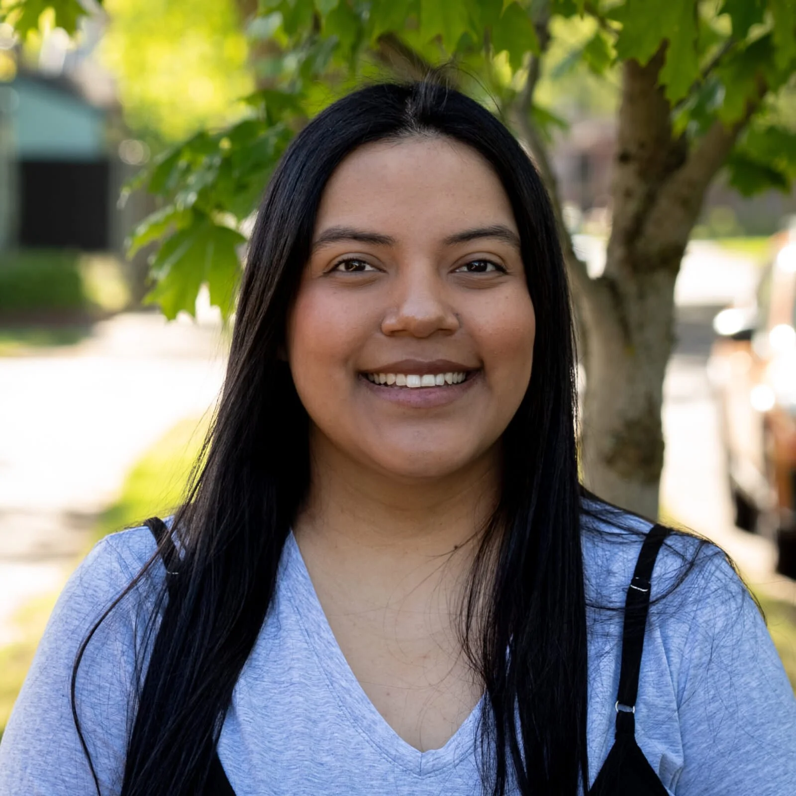 Close-up of a smiling woman with long black hair, outdoors near a tree, with a blurred background of greenery and parked cars.