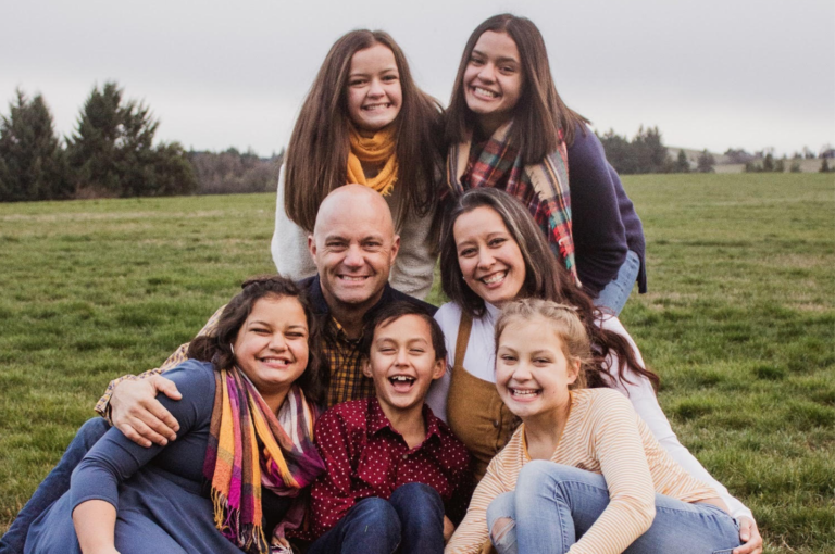 A happy family with three adults and four children smiling outdoors in a grassy field.