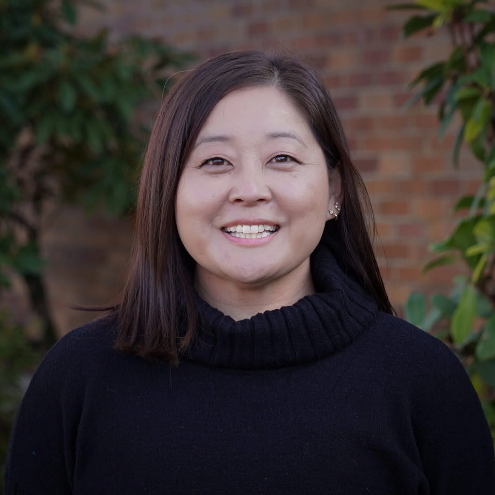 A woman with shoulder-length dark hair smiling outdoors in front of a brick wall and green foliage, wearing a black turtleneck sweater.