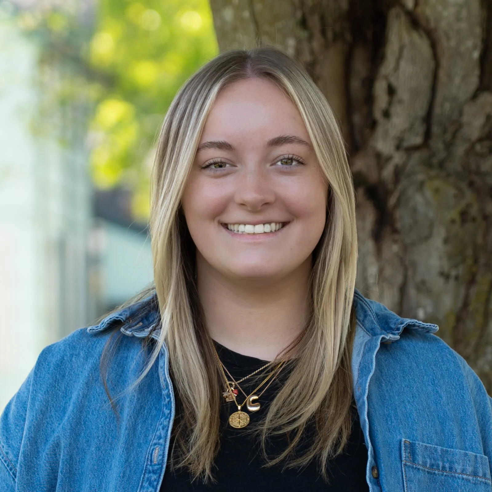 A young woman with blonde hair smiling outdoors, wearing a black shirt, a blue denim jacket, and layered gold necklaces, standing in front of a tree with green leaves and sunlight filtering through.