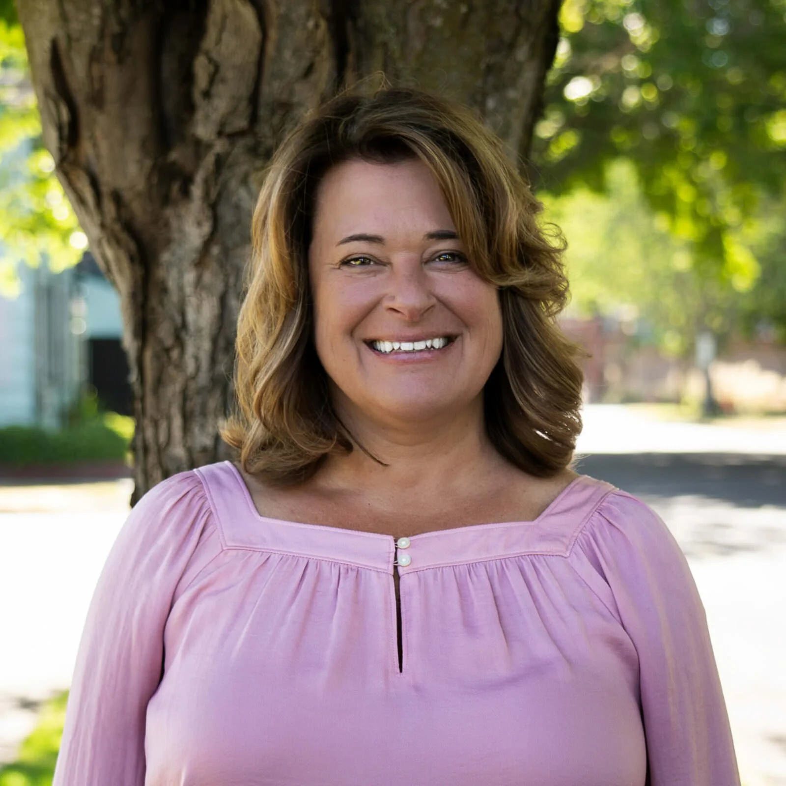 A woman smiling outdoors in front of a tree, wearing a white sleeveless top.