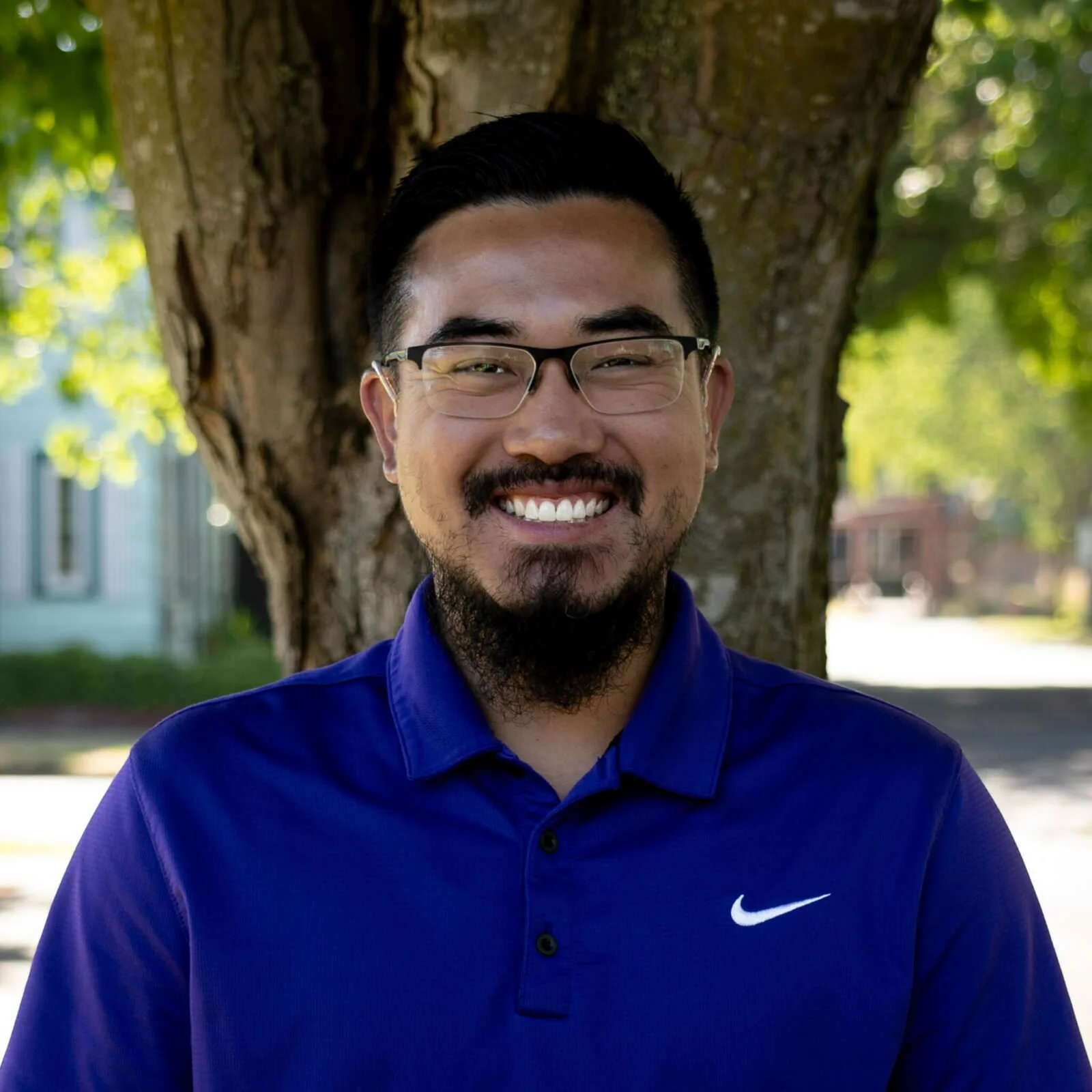 A smiling man with glasses and a beard, wearing a blue Nike polo shirt, standing outdoors in front of a tree with green leaves and a background of houses and street.