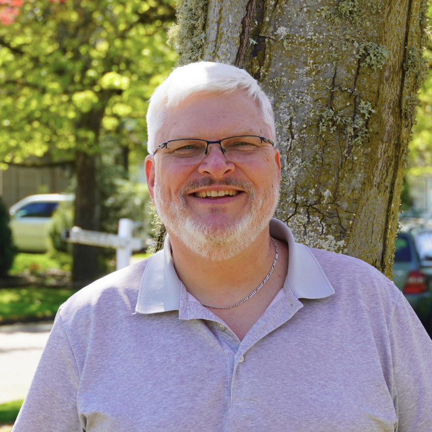 A middle-aged man with gray hair, glasses, and a beard, smiling outdoors next to a tree with a blurred background of green foliage.