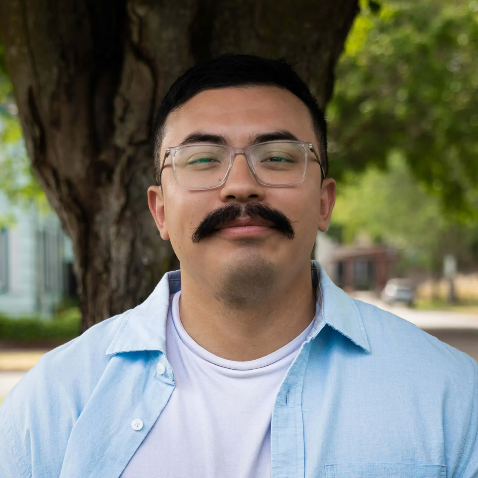Man with glasses, mustache, and light blue shirt standing outdoors near a large tree with green leaves in the background.