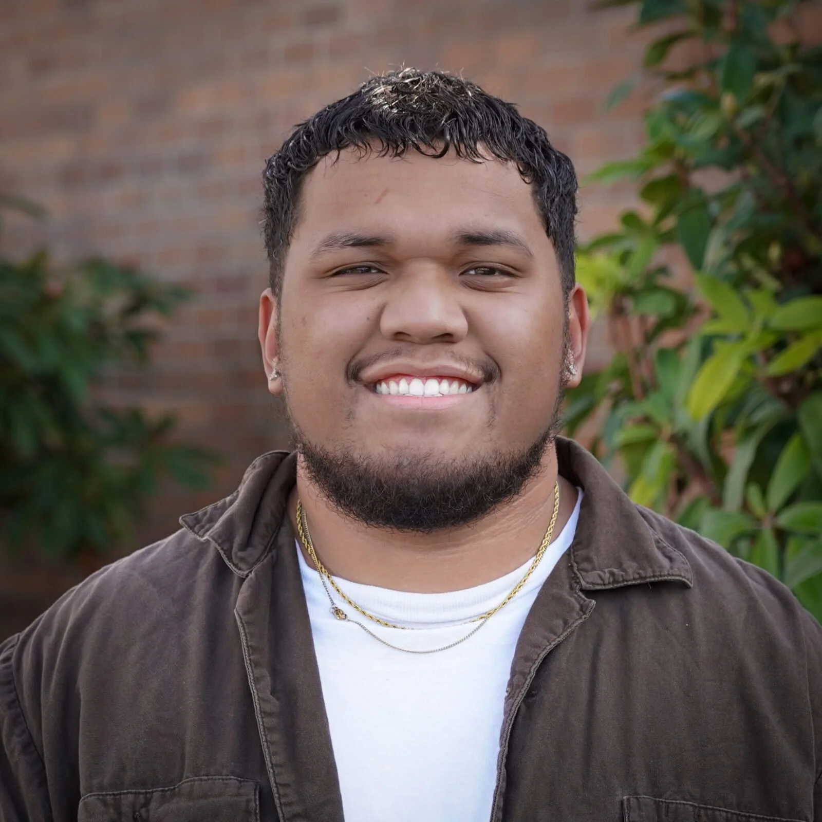 A young man smiling outdoors with green plants and a brick wall in the background.