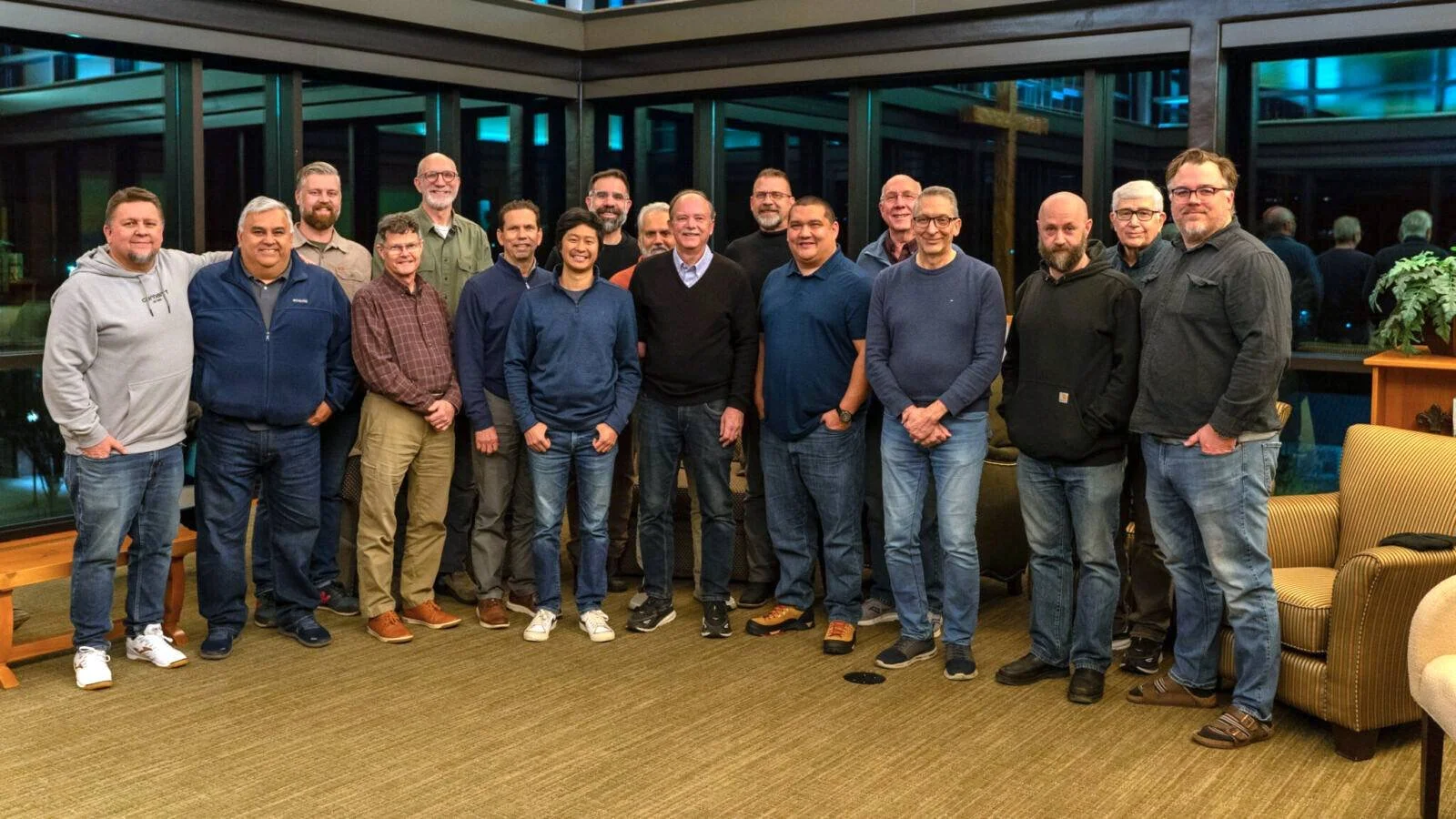 A group of 17 people posing for a photo indoors, standing in front of large glass windows at night, with reflections of interior lights and some greenery outside.