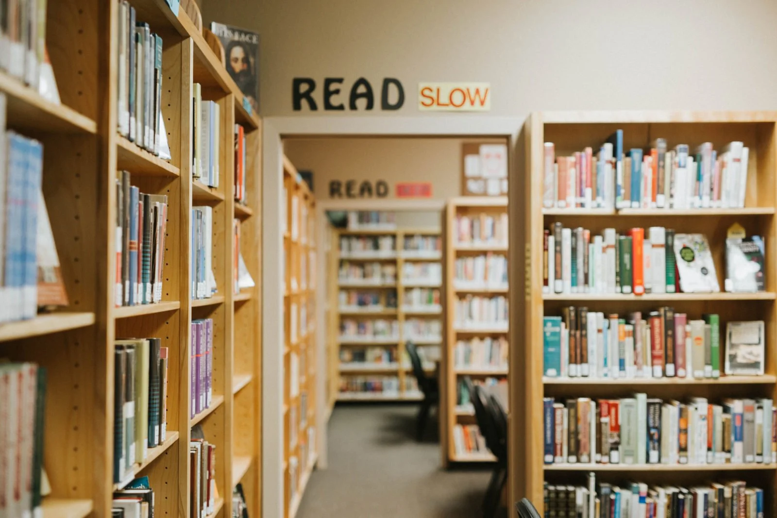 A view inside a library with wooden bookshelves filled with books. The hallway continues with more shelves and chairs in the background.
