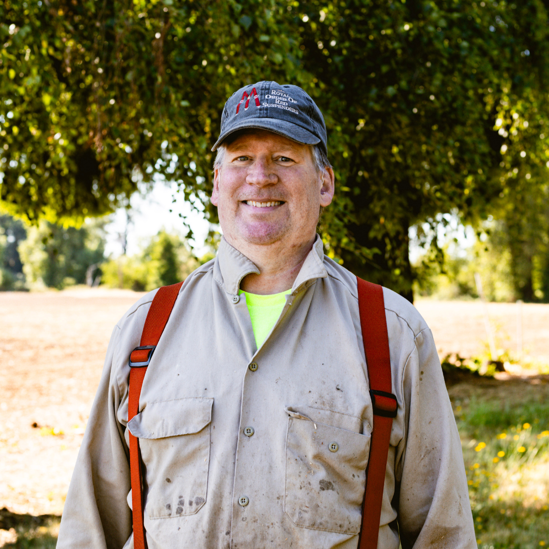 Smiling man outdoors wearing a baseball cap and beige work jacket with suspenders, standing in front of a large tree on a sunny day.