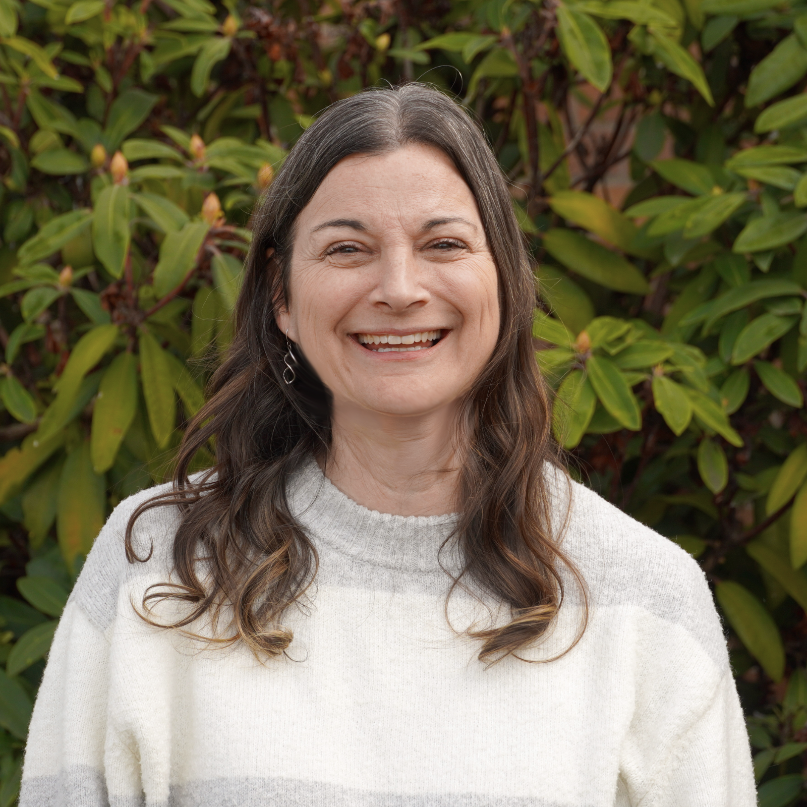 Smiling woman with long wavy brown hair and earrings, wearing a white sweater, standing outdoors in front of green bushes.