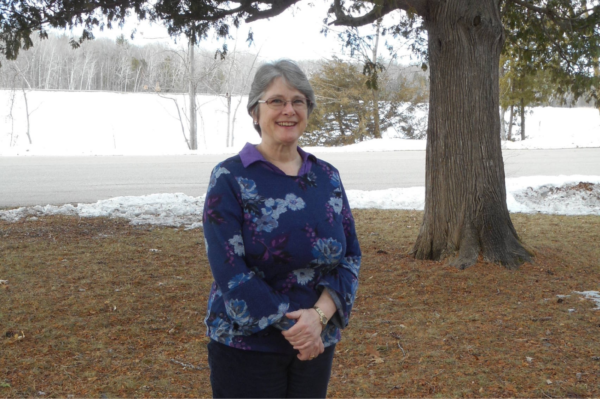 A woman with gray hair and glasses standing outdoors near a large tree with snow on the ground and a road in the background.