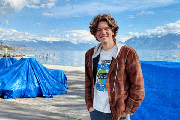 A young man with curly hair stands outdoors near a large lake with mountains in the background. He is wearing a brown jacket over a white T-shirt and is smiling.