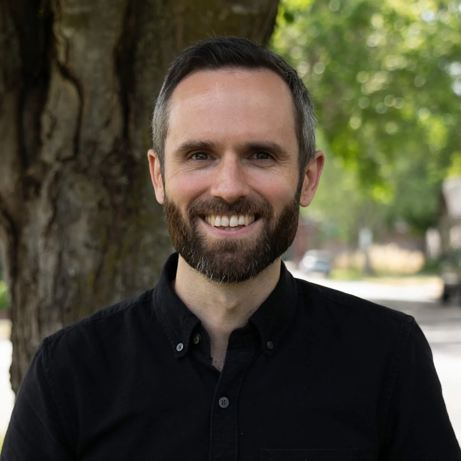 Man with short dark hair and beard smiling outdoors in front of a large tree, wearing a black button-up shirt.