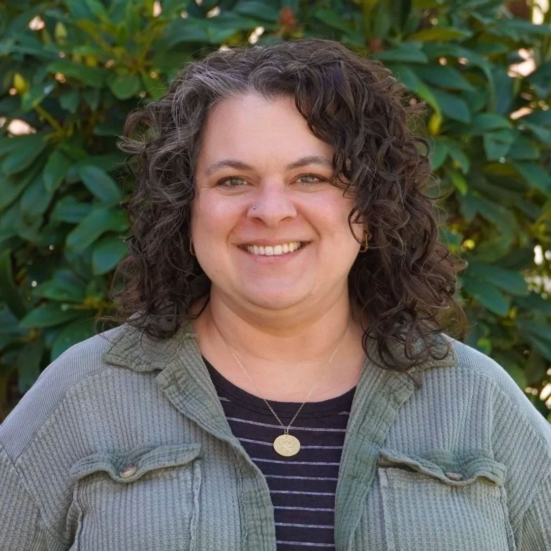 A woman with shoulder-length dark curly hair smiling, wearing large gold earrings, a small nose piercing, a necklace with a small pendant, and a black and white striped sleeveless top, standing outdoors in front of a tree with sunlight filtering through the leaves.