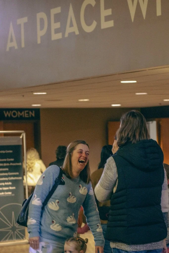 Two women laughing and talking in a public indoor space, with one woman holding a small child, in front of a sign for the women's restroom.