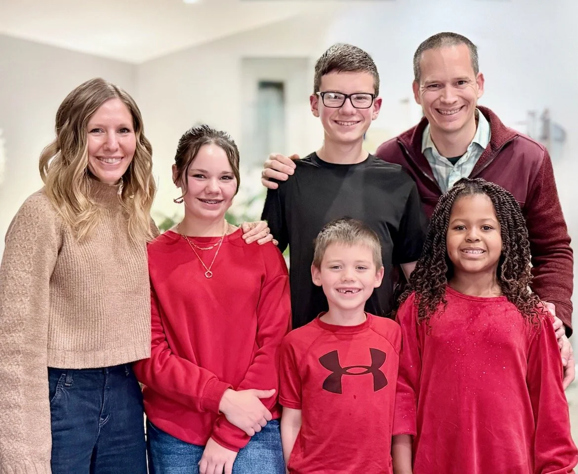 A family of seven, including two adult women, two adult men, and three children, smiling and standing indoors in a warmly lit room, casually dressed.