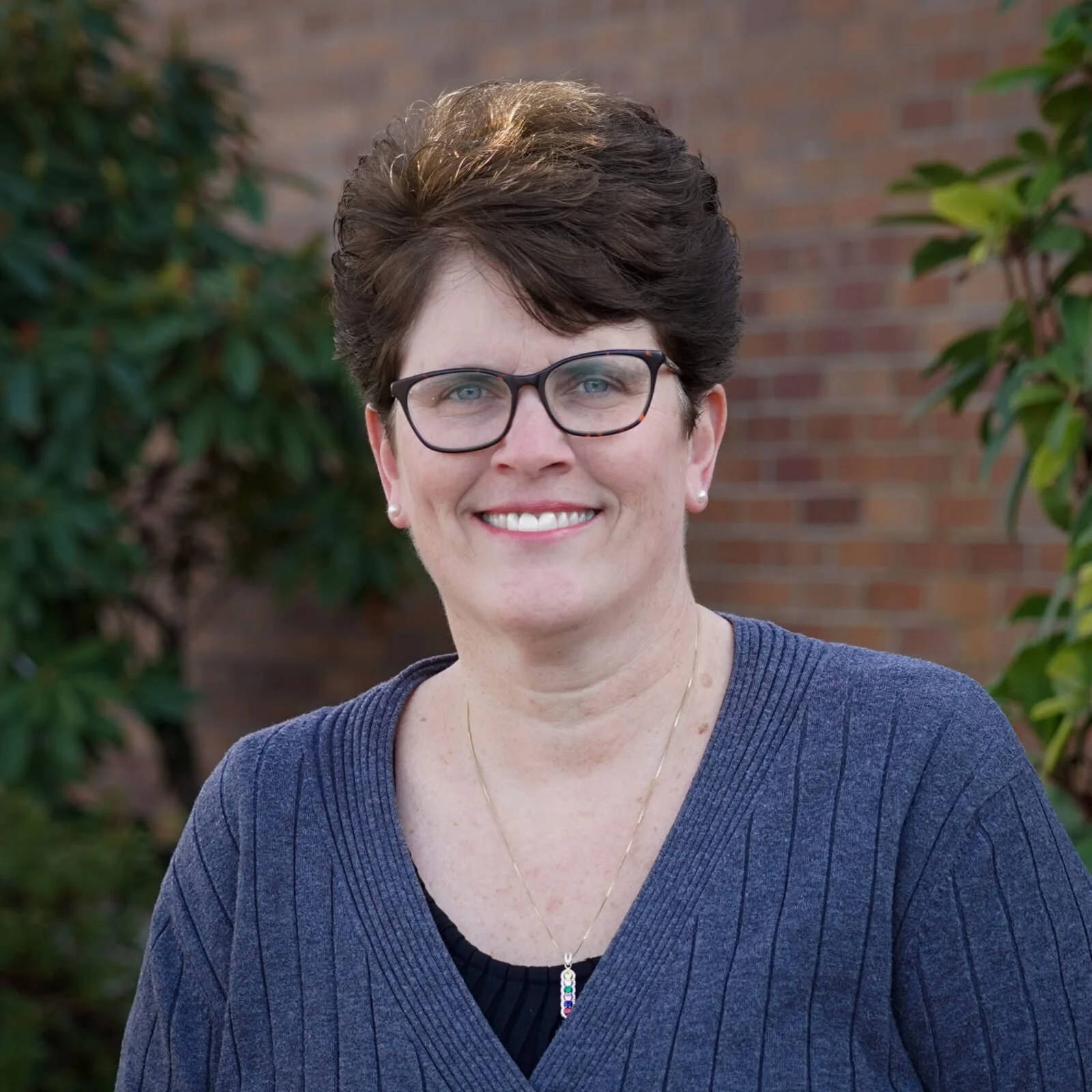 A woman with short dark hair, glasses, and a blue sweater standing outdoors with a brick wall and green plants in the background.