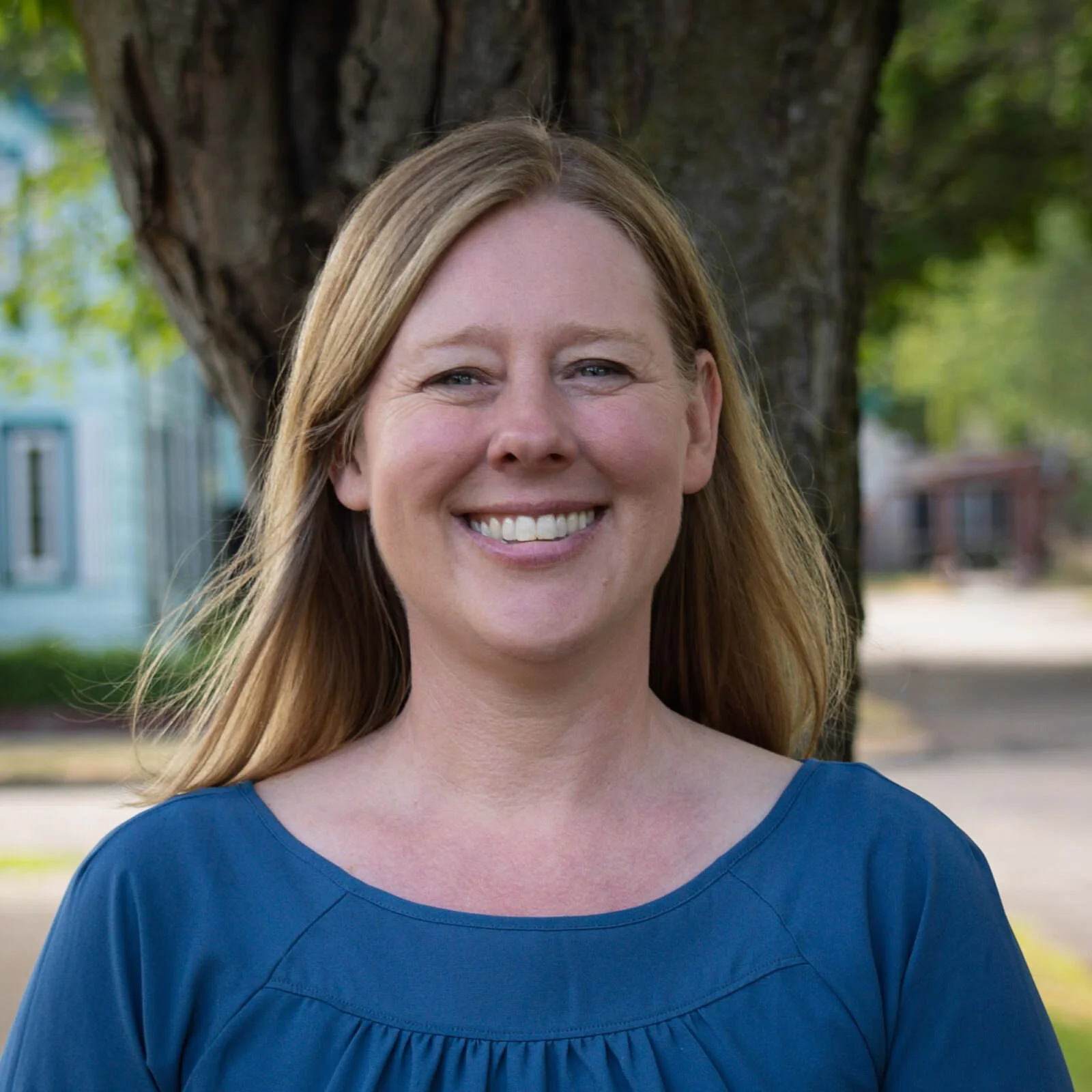 A smiling woman with light brown hair wearing a blue top standing outdoors in front of a tree with a blurry background of buildings and greenery.