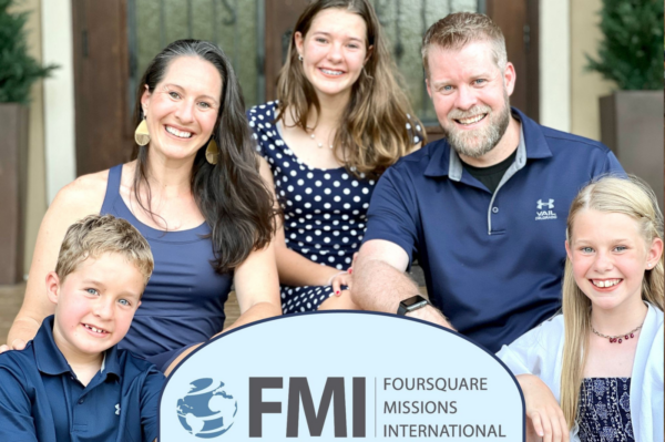 A family of five smiling outdoors, holding a sign with the logo of Foursquare Missions International.