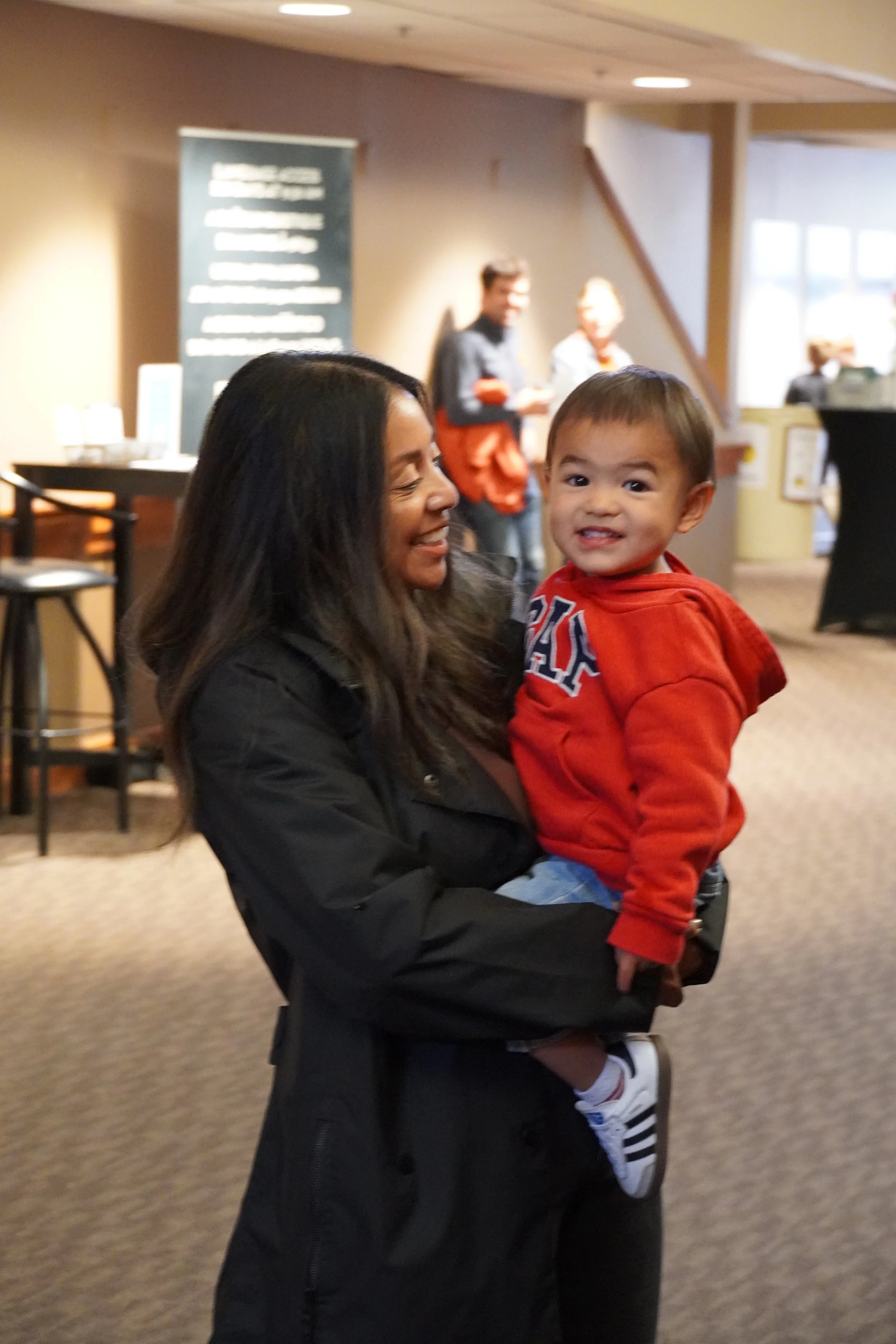 A woman holding a young boy in a red hoodie, smiling in an indoor setting with people in the background.