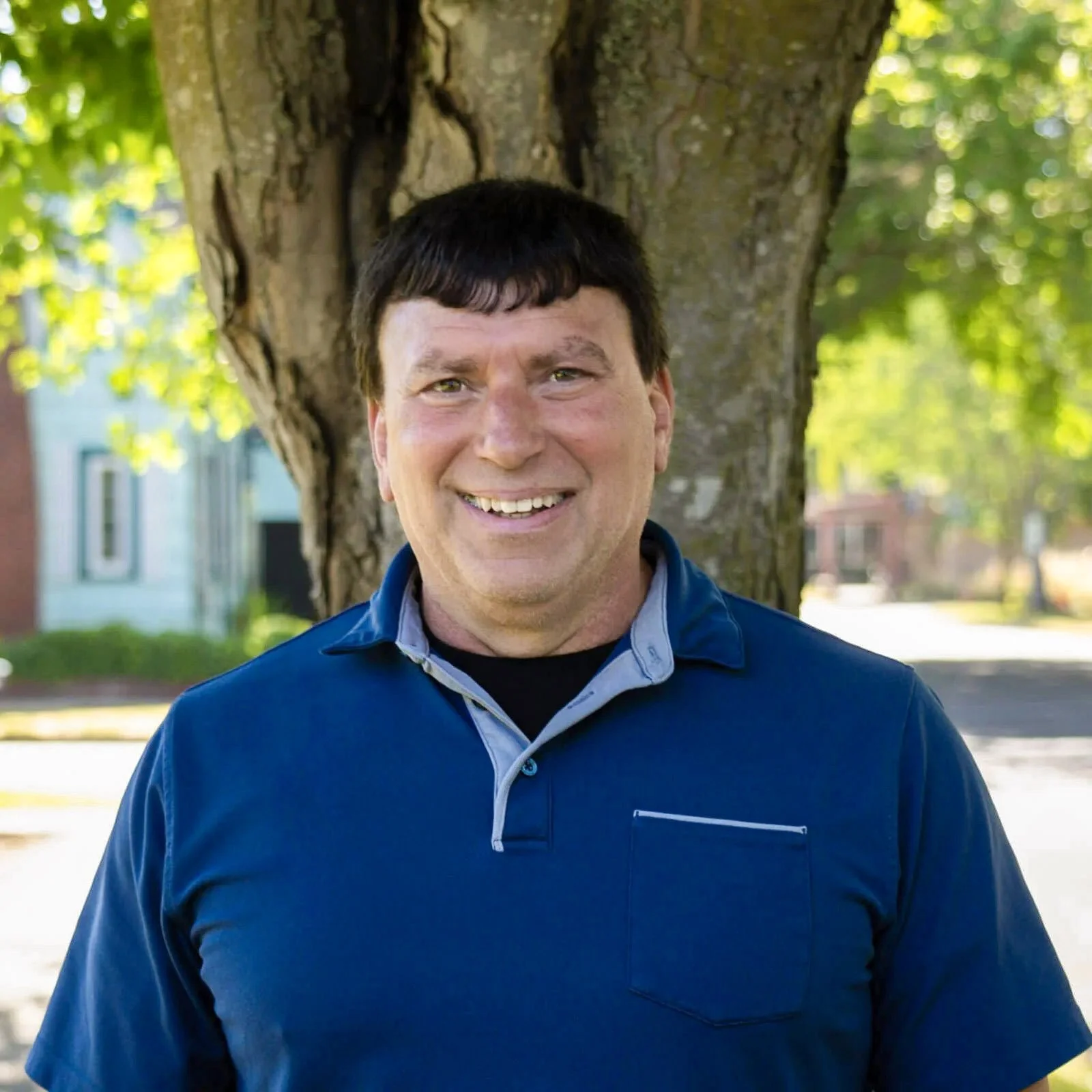 A man smiling outdoors standing in front of a tree with green leaves, wearing a blue collared shirt.