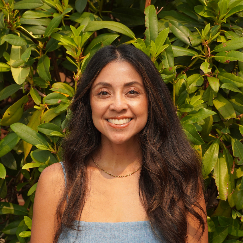 A woman smiling outdoors in front of a tree, wearing a white sleeveless top.
