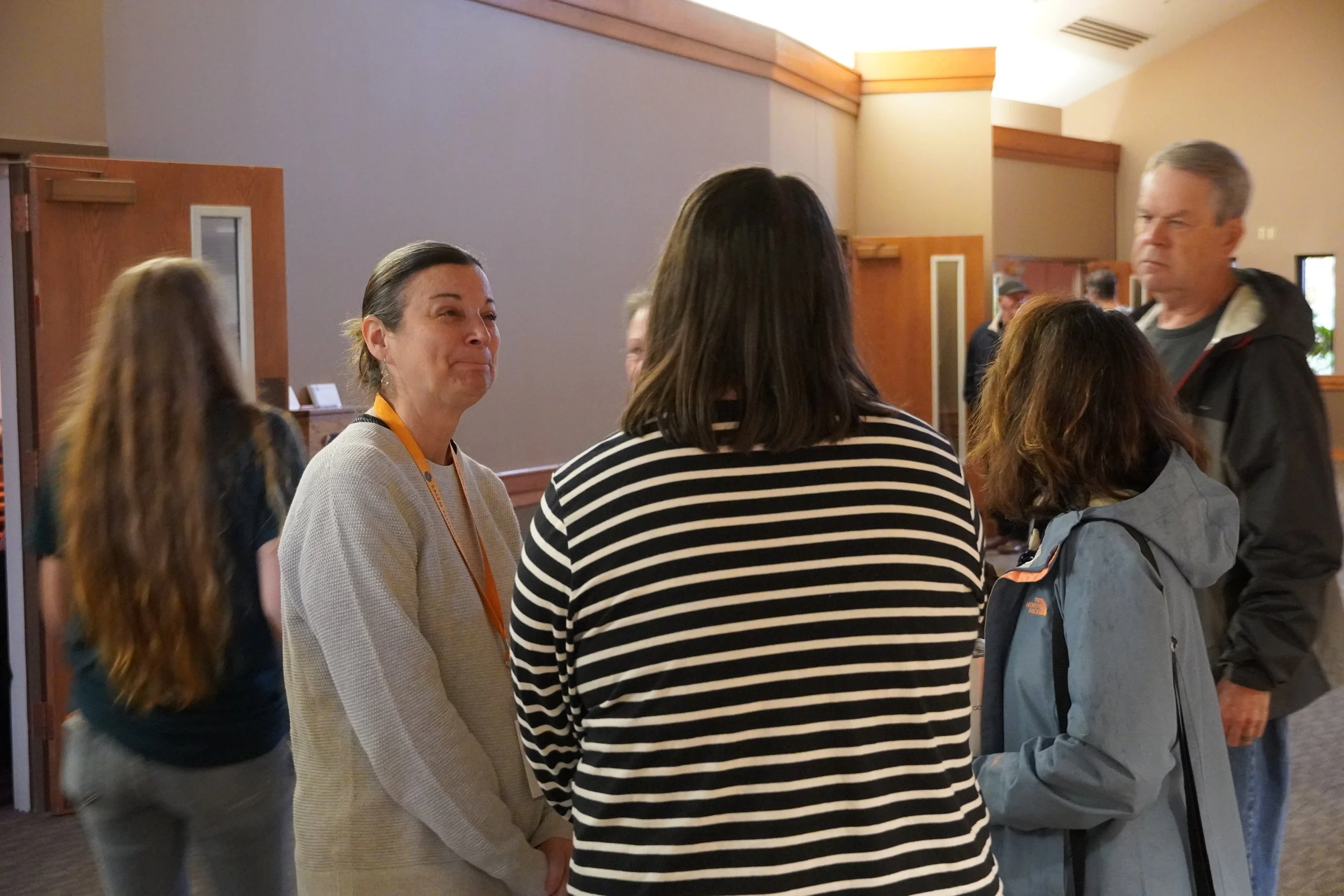 Group of people gathered indoors, engaging in conversation, with wooden accents and neutral-colored walls in the background.