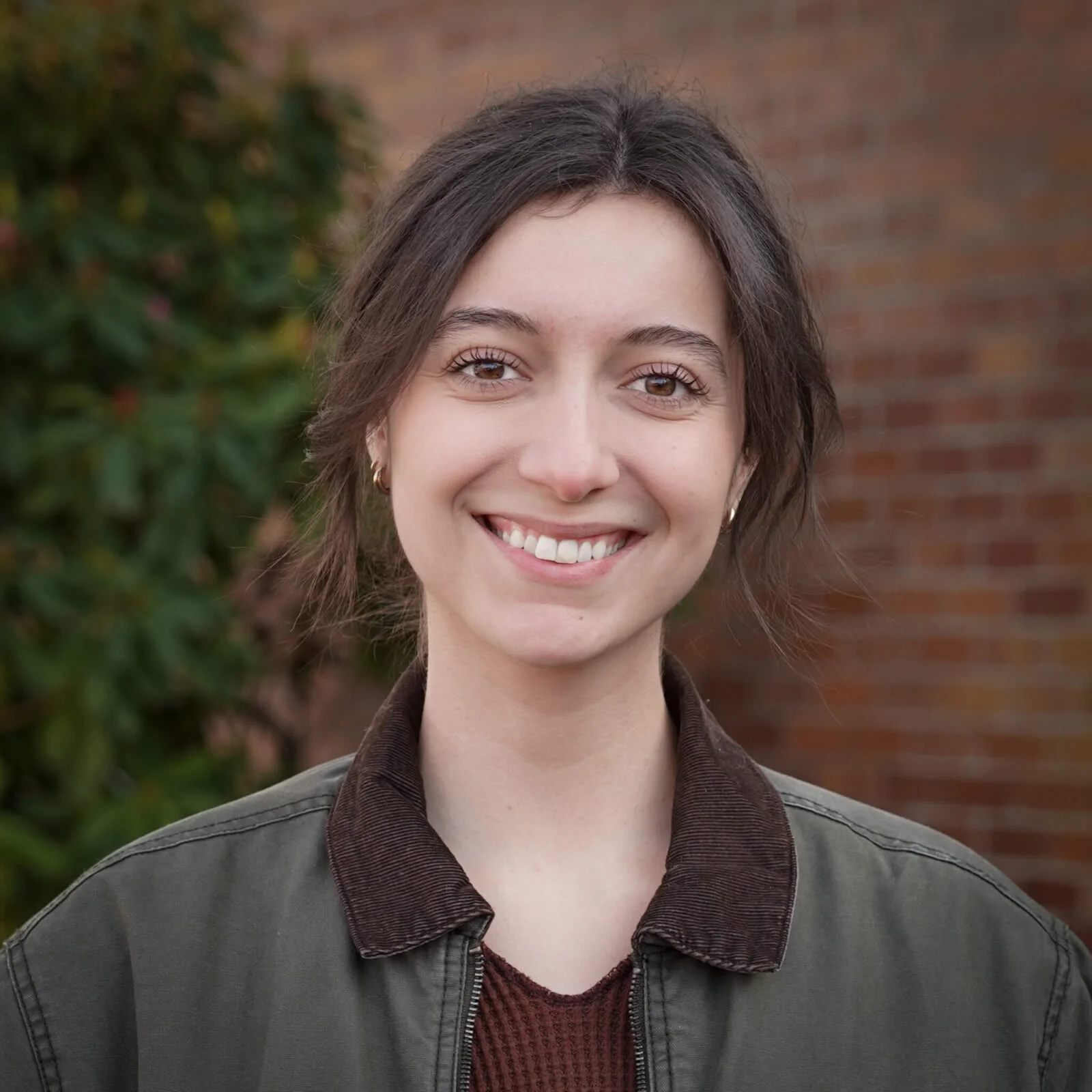 A young woman with short brown hair smiling outdoors, wearing a dark jacket and a maroon top, with greenery and a brick wall in the background.