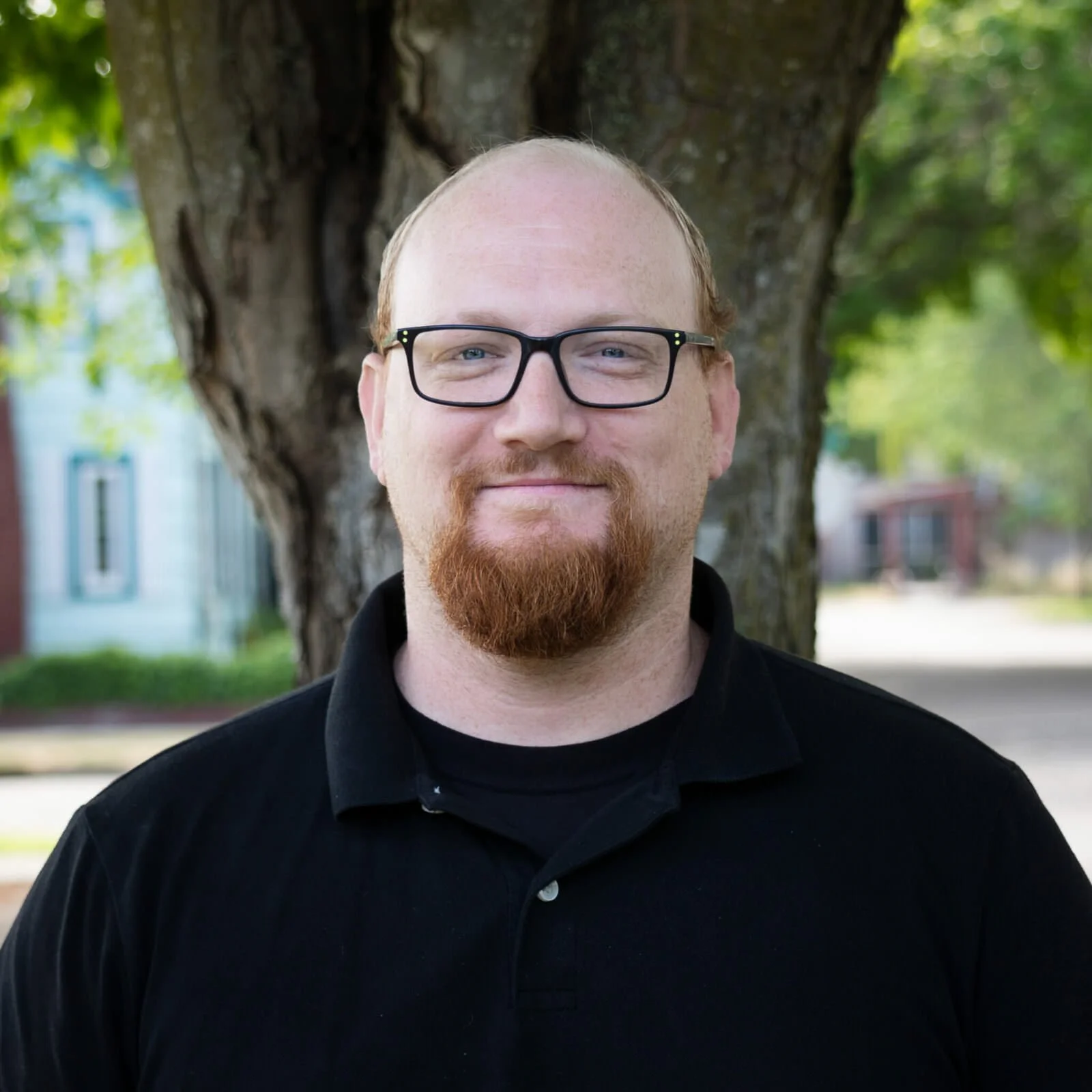A man with glasses, a red beard, and a black shirt standing outdoors in front of a large tree.