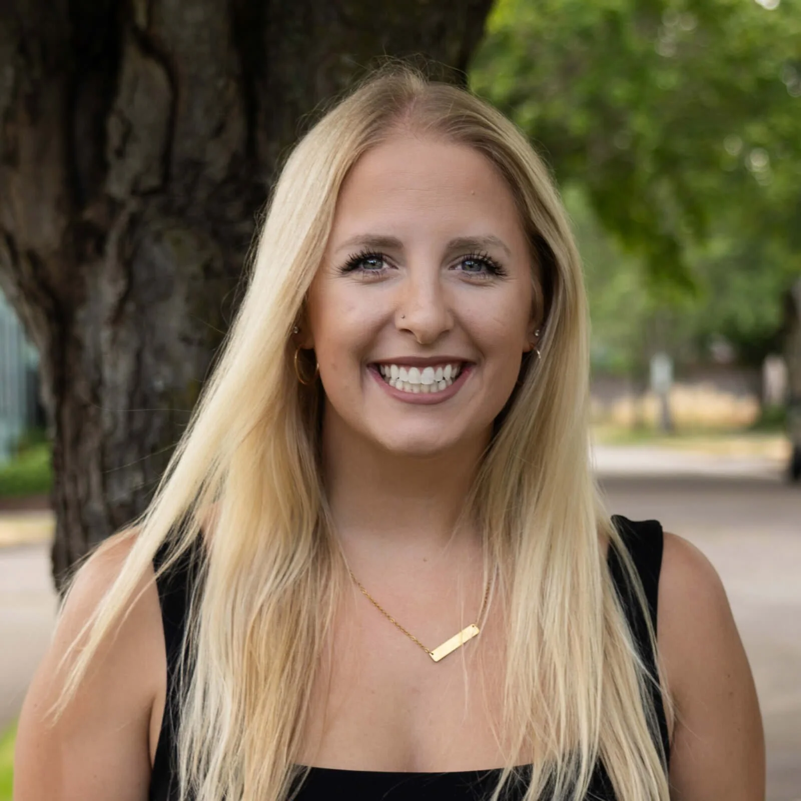A woman with long blonde hair and a black sleeveless top standing outdoors in front of a large tree with green foliage, smiling at the camera.