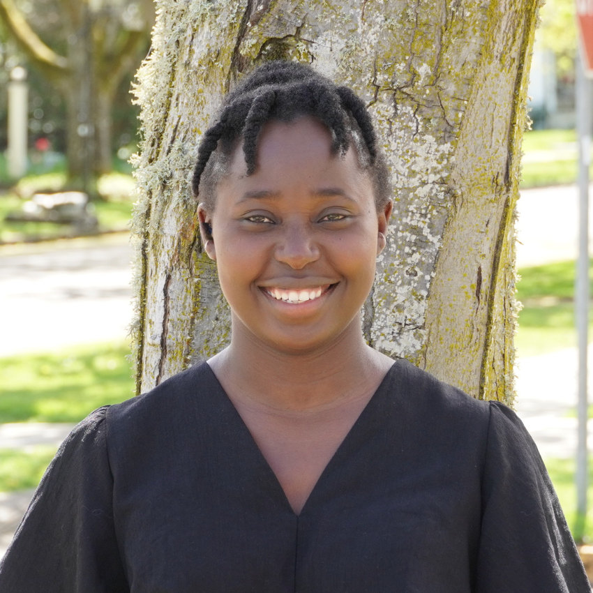 Young woman with styled hair, standing outdoors with trees and buildings in the background, smiling.