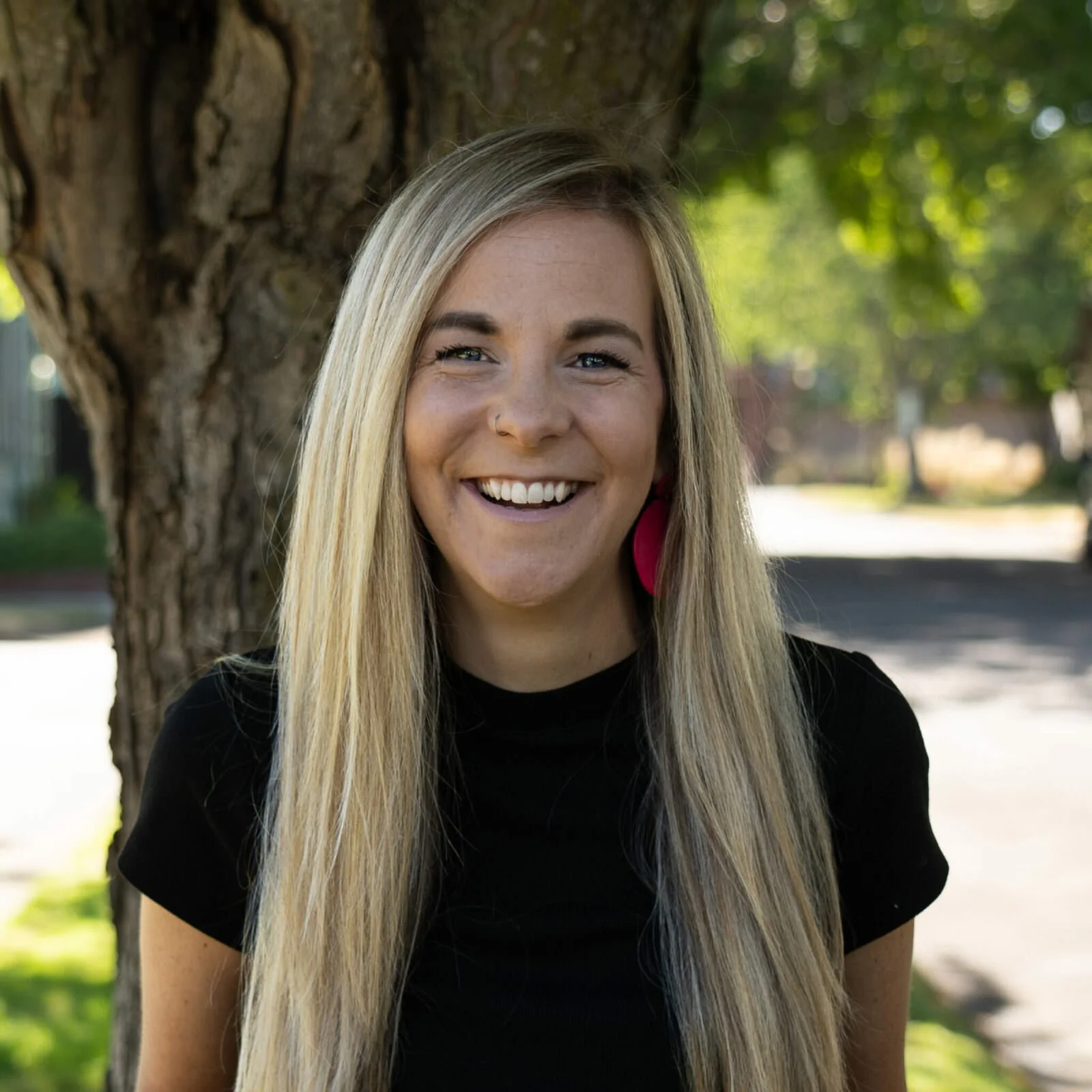 A woman with long blonde hair smiling outdoors, standing next to a tree with green leaves in the background.