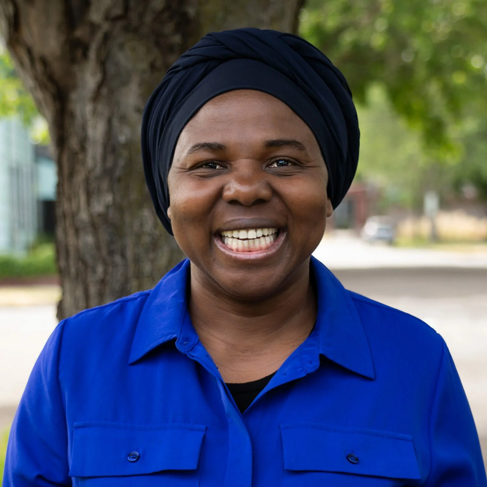 A woman with a dark blue headwrap and matching blue shirt smiling outdoors near a tree.