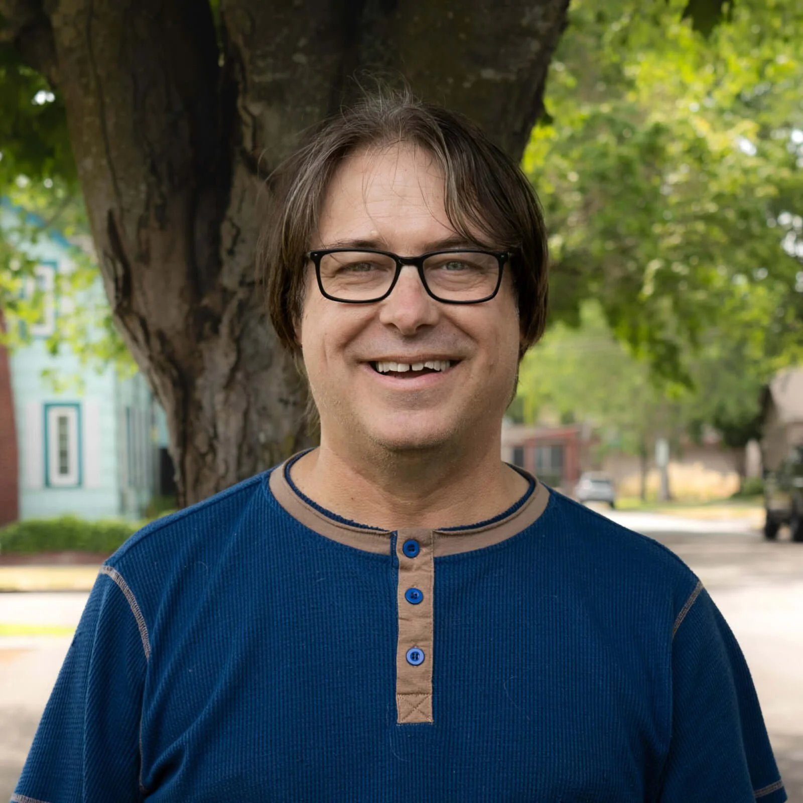 A man smiling outdoors in front of a tree, wearing glasses and a blue shirt.