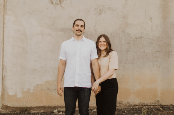 A man and woman holding hands and smiling while standing outdoors against a weathered beige wall.