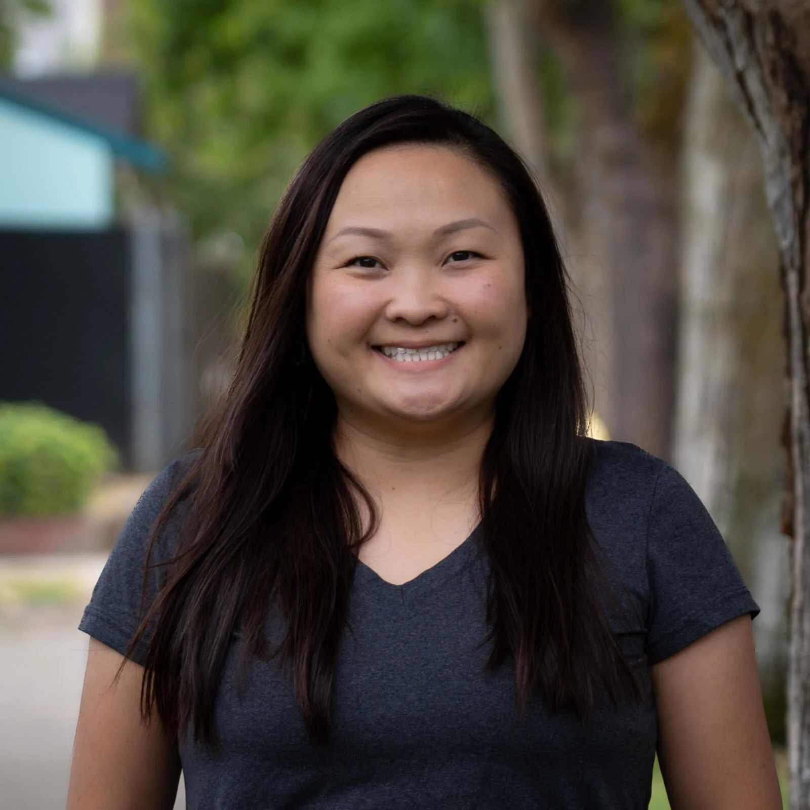 A smiling woman with long dark hair, wearing a dark gray T-shirt, standing outdoors with trees and a building background.