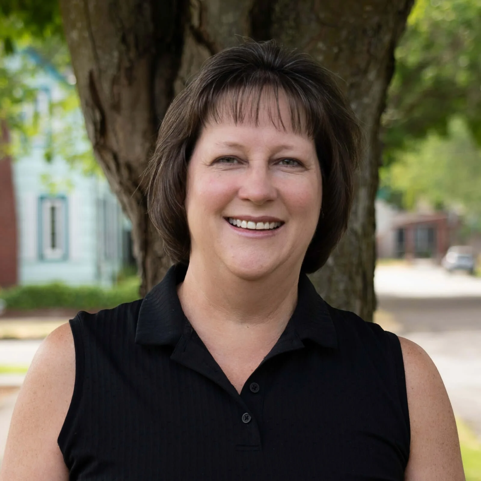 A woman with short dark brown hair smiling outdoors in front of a tree with a blurred background of houses and greenery.