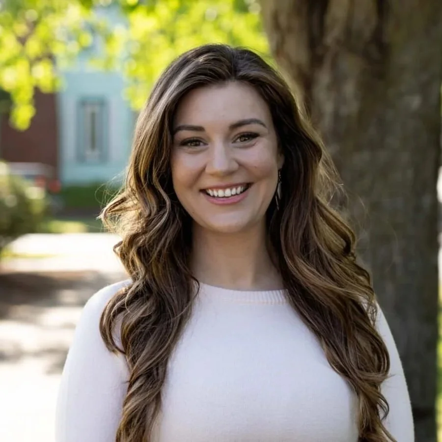 A young woman with long, wavy brown hair smiling outdoors on a sunny day, wearing a white top.
