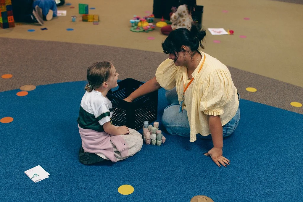 A young girl sitting on a blue carpet with a woman kneeling beside her, playing with toys in a colorful playroom.