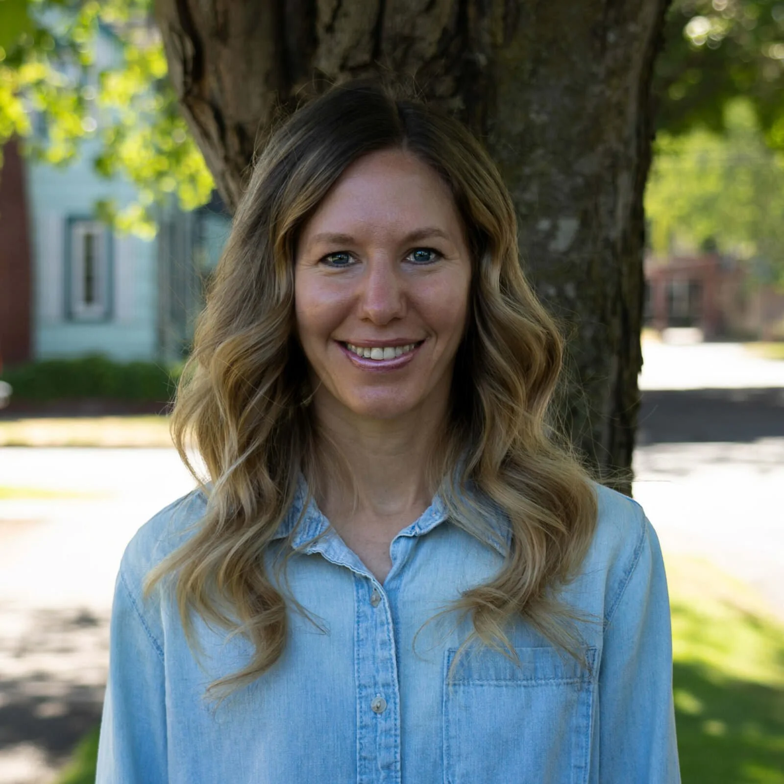 A woman with wavy blonde hair standing outdoors in front of a large tree, smiling, wearing a light blue denim shirt.