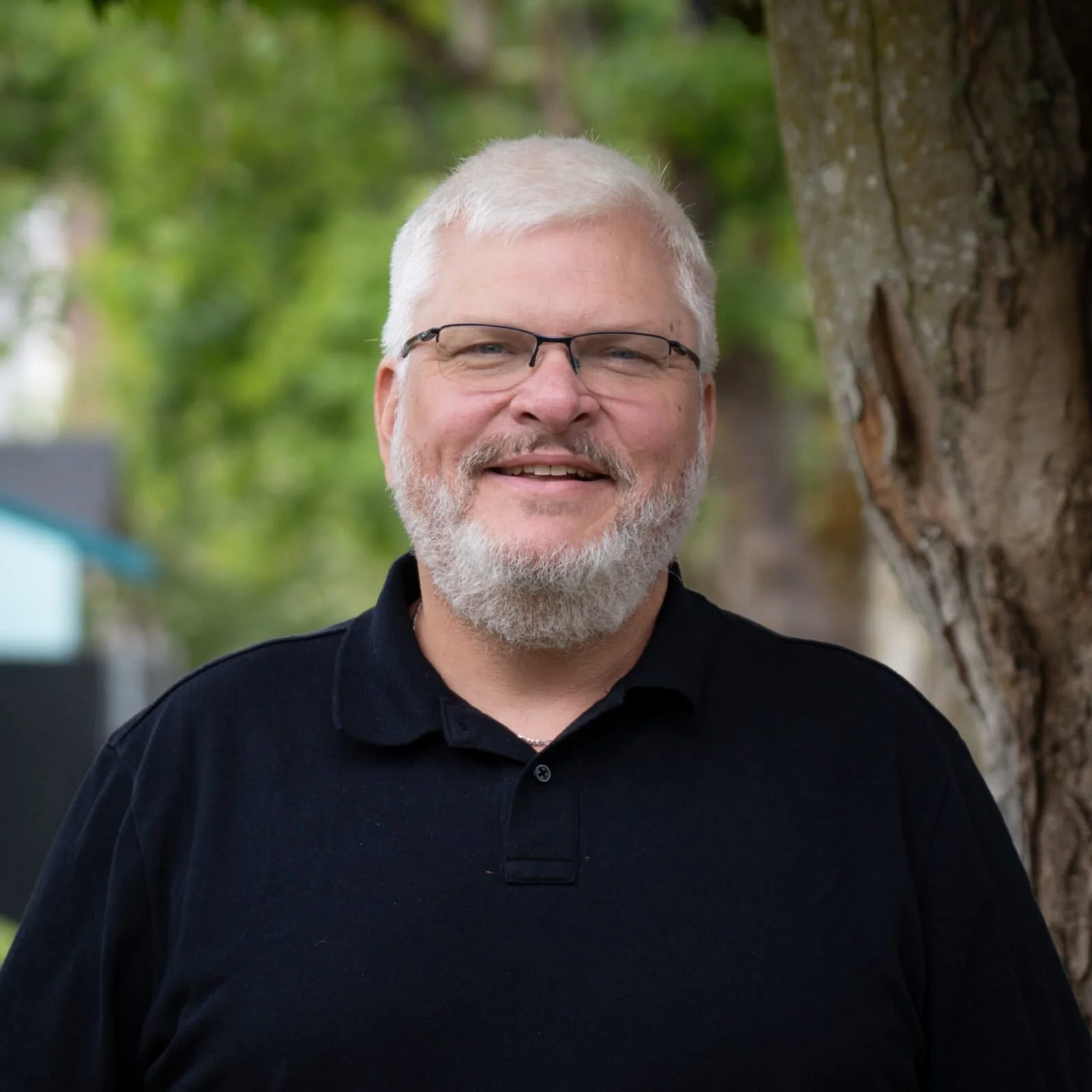 A middle-aged man with gray hair, glasses, and a beard, smiling outdoors next to a tree with a blurred background of green foliage.