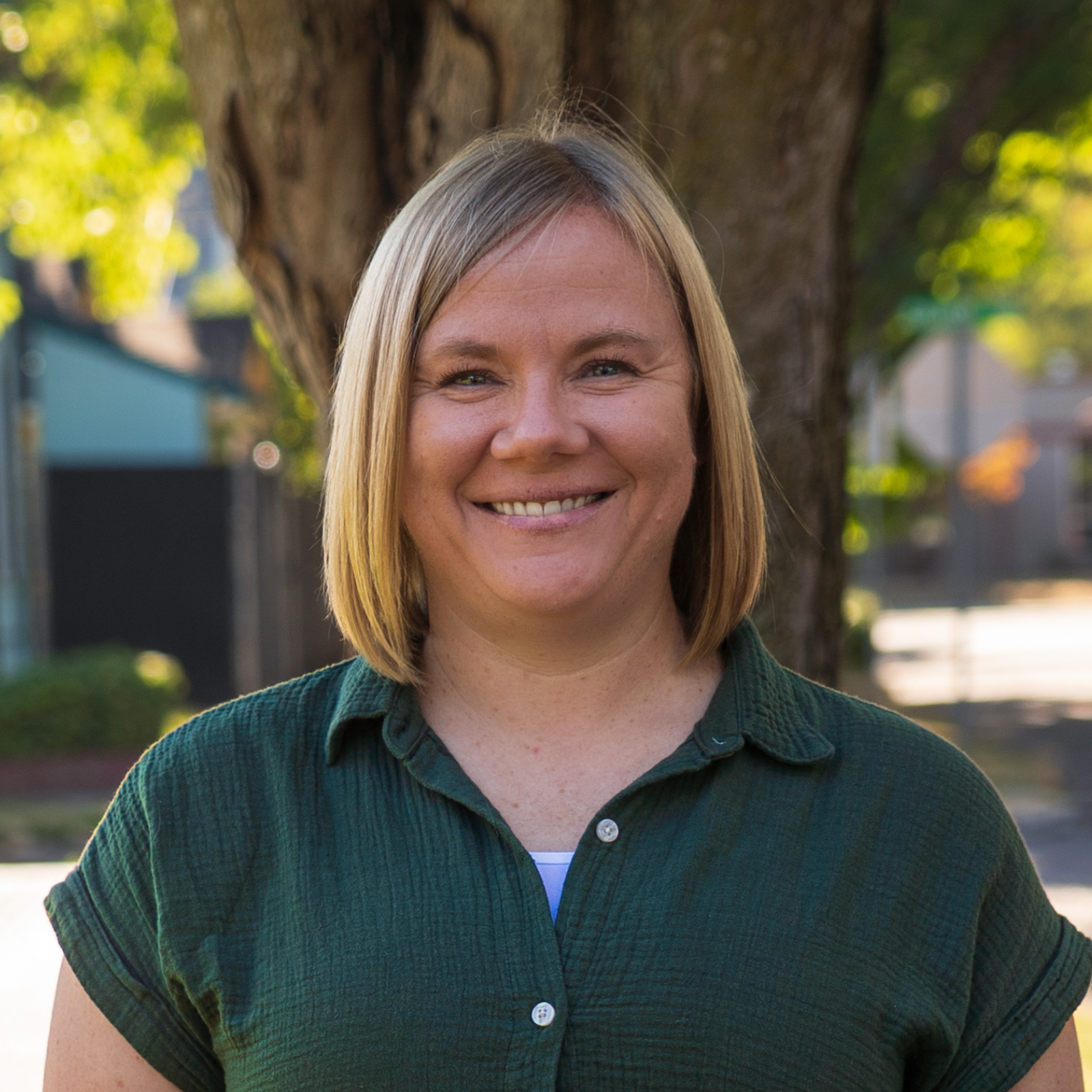 A woman with shoulder-length blonde hair smiling outdoors in front of a tree with green leaves and a residential neighborhood in the background.