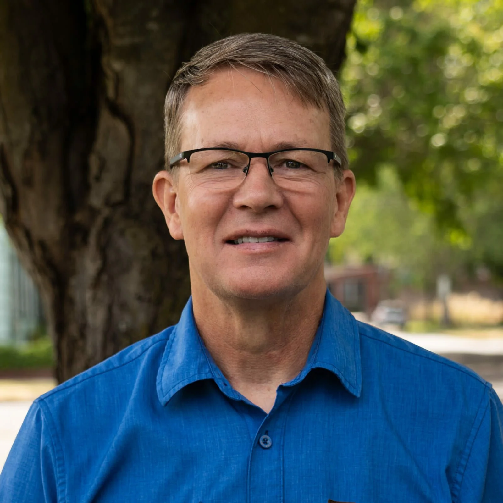 A man wearing glasses and a blue button-up shirt, standing outdoors in front of a large tree and greenery, smiling at the camera.