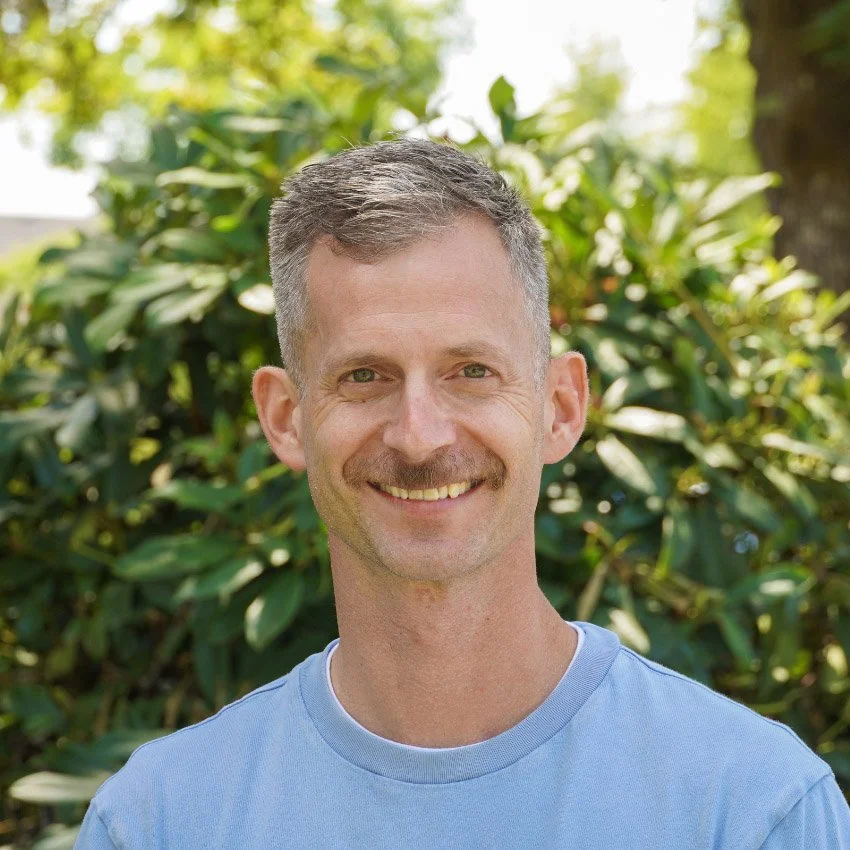A man with short dark hair, a mustache, and a blue button-up shirt standing outdoors in front of a tree, smiling at the camera.