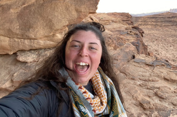 A woman smiling and taking a selfie outdoors in a rocky desert landscape.