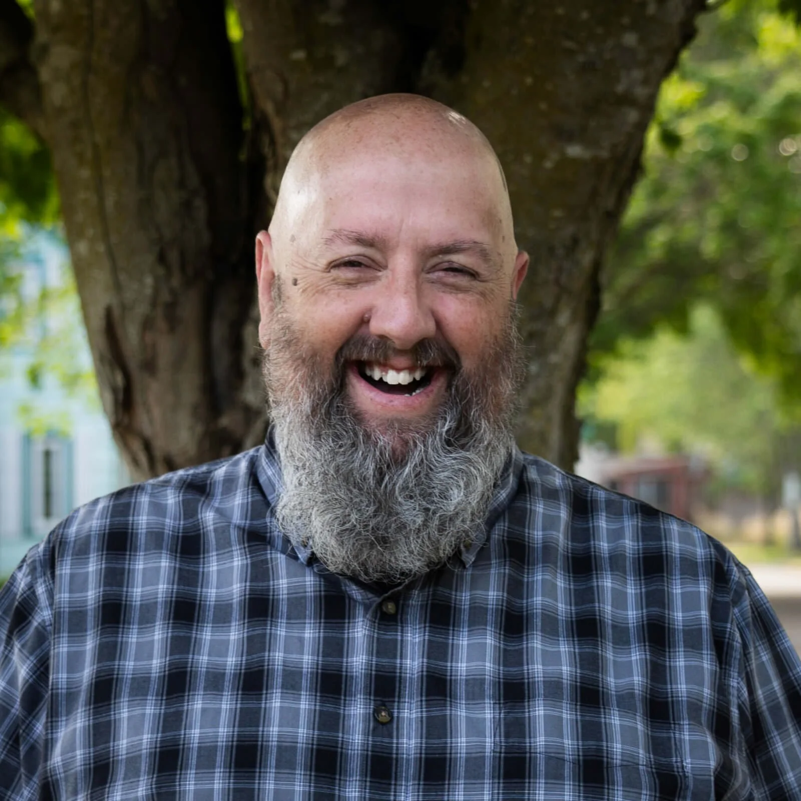 A smiling man with a gray beard and bald head standing outdoors in front of a tree, wearing a blue plaid shirt.