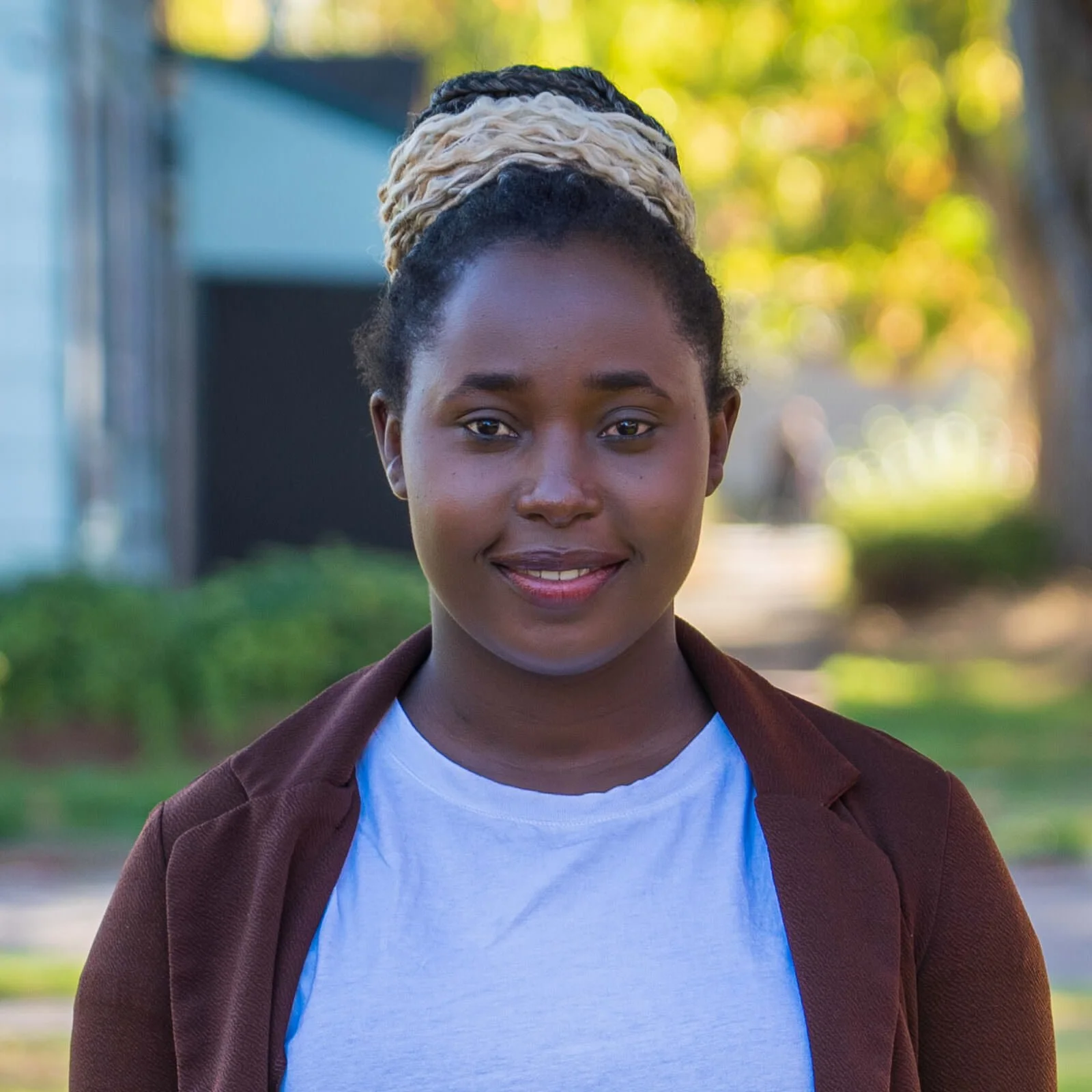 Young woman with styled hair, standing outdoors with trees and buildings in the background, smiling.
